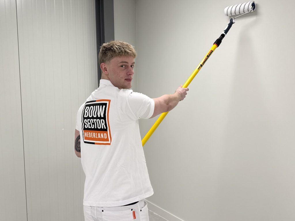 Jonge schilder kijkt om terwijl hij met een gele verlengsteel een witte muur saust