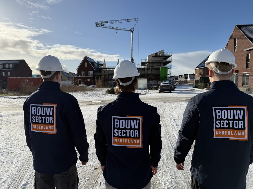 Drie vakmensen van Bouwsector Nederland met witte helm kijken naar nieuwbouwhuizen en een bouwkraan in de sneeuw