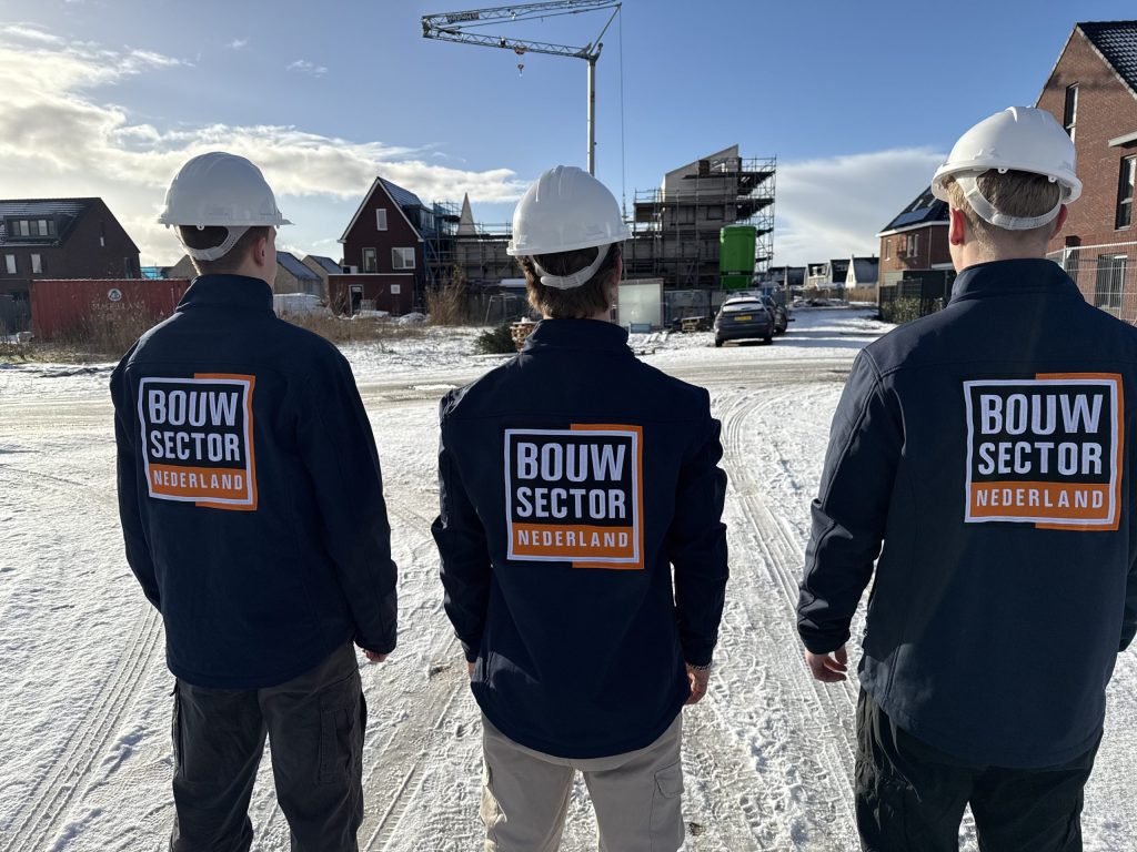 Drie vakmensen van Bouwsector Nederland met bouwhelm, van achteren gezien, kijken naar een straat met nieuwe huizen en een bouwkraan