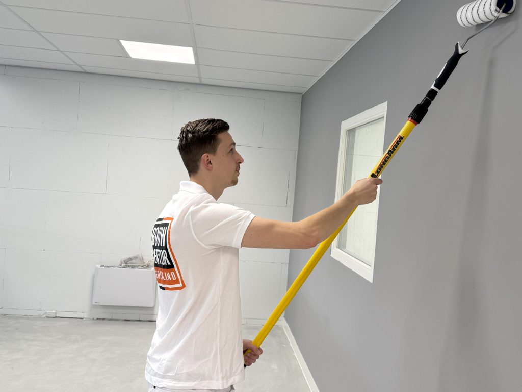 Schilder zet een grijze wand naast een raam met de roller aan een lange steel, achter hem een nog kale wand